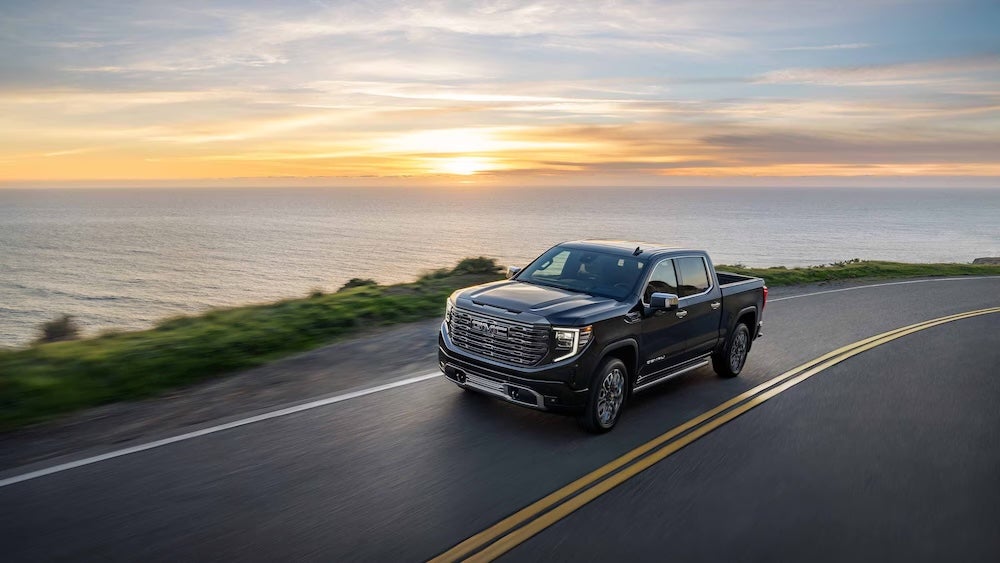 A black GMC pickup truck driving down a coastal road at sunrise